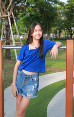 Fototapeta premium Portrait of young girl in blue shirt is leaning against the playground bars, while looking at camera.