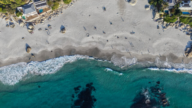 Aerial view of Clifton 1st beach with white sand, turquoise ocean waves, large granite boulders, and people relaxing on the shore in Cape Town, Western Cape, South Africa.