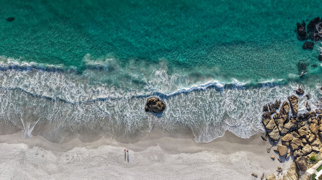 Aerial view of Clifton 1st beach with turquoise waves, a large rock, and two people walking on the white sand in Cape Town, Western Cape, South Africa.