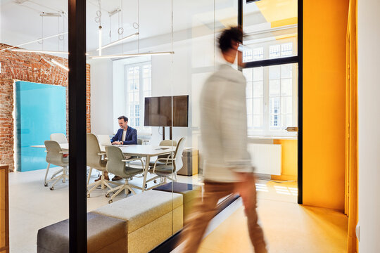 Businessman works in office while male colleague walks past