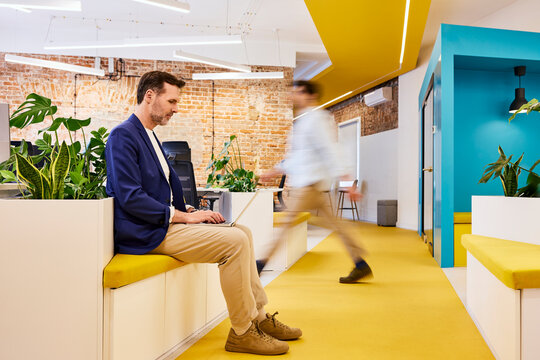 Businessman uses laptop in modern office with walking colleague