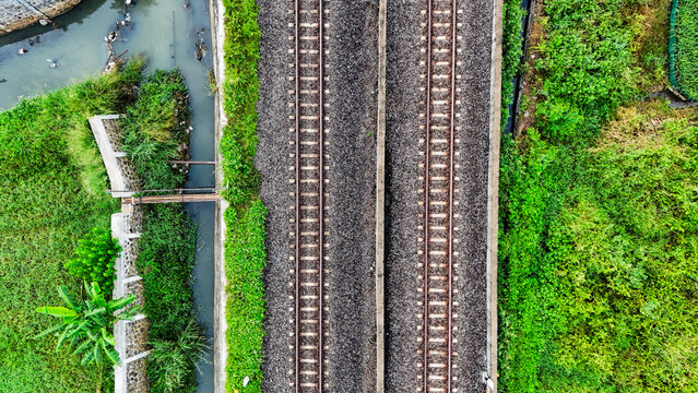Aerial view of the railway tracks in Gedebage, surrounded by lush green vegetation and a small canal in Bandung, West Java, Indonesia.