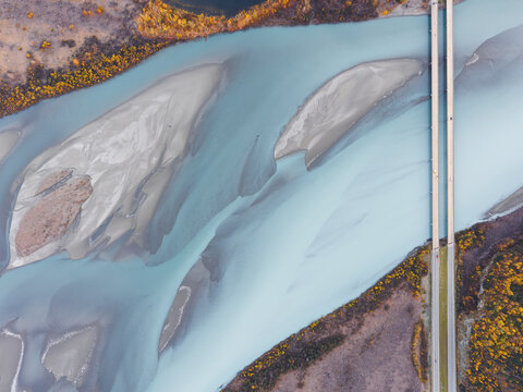 Aerial view of the Knik River with its glacial blue water, intricate sandbars, and a bridge crossing the landscape surrounded by autumn foliage in Knik River, Alaska, United States.