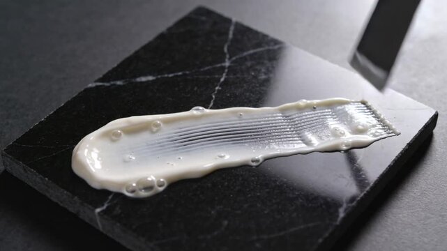 Entering top-right, metal spatula dragging white smear across black marble tile, for smoothing