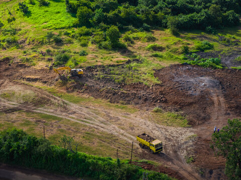 Aerial view of the Athani Sugars Limited Rayat Unit construction site with an excavator and yellow dump truck on dirt tracks surrounded by green vegetation in Shevalwadi, Maharashtra, India.