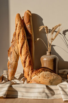 Various baguettes and sourdough bread loaf on linen tablecloth, vase with wheat spikelet, empty beige neutral wall background with natural warm sunlight shadows, cottage home interior, rustic bakery