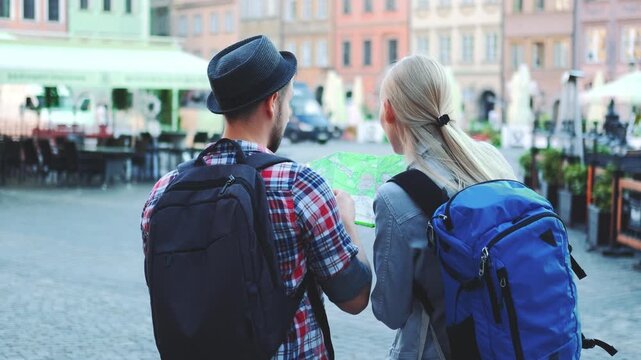 A man and woman tourists stand together in a European city square during daytime, examining a large map to plan their next destination.