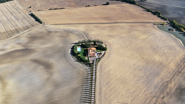 Aerial view of a secluded farmhouse with a swimming pool and a cypress-lined driveway surrounded by rolling golden fields in Castiglione d'Orcia, Tuscany, Italy.
