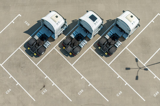 Aerial view of three white semi-truck cabs parked in a marked parking lot with visible parking space numbers and shadows cast on the asphalt in Warszawa, Masovian Voivodeship, Poland.