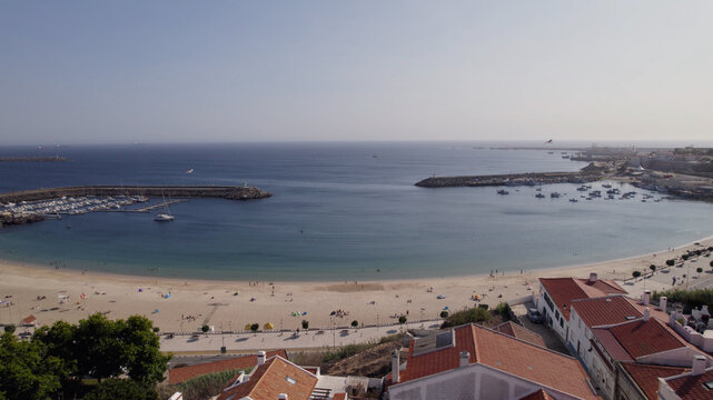 Aerial view of the sandy beach and harbor in the coastal town of Sines with red-tiled rooftops and calm blue waters in Sines, Setubal, Portugal.