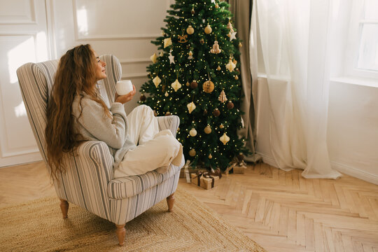 Young beautiful woman drinking tea sitting under decorated Christmas tree.