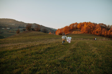 Naklejka premium Happy girl running with outstretched arms and pink and white balloons across a green autumn meadow.