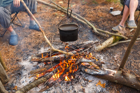 Metal pot hanging over campfire with burning logs surrounded by tourists sitting around
