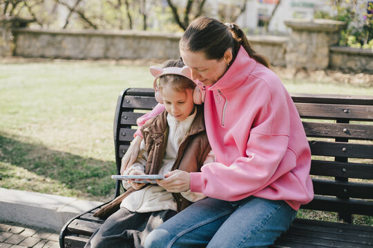 Happy mother and daughter sitting on a park bench watching tablet.
