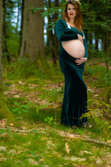 Side View of Expectant Red Haired Mother in an Elegant Green Dress in the Forest. A pregnant red-haired woman poses in profile in a quiet woodland, wearing an elegant long dark green dress and a wreat © Nadiia