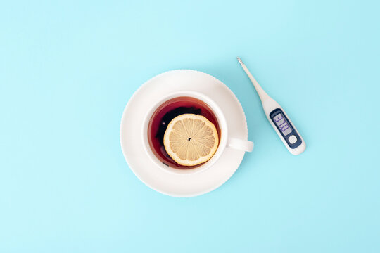 Digital thermometer beside hot tea with lemon on a bright blue background, illustrating fever monitoring, illness care and seasonal health. Top view, flat lay