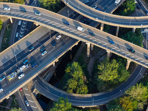 Aerial view of a complex highway interchange with multiple levels and cars driving on roads surrounded by lush green trees in the United Kingdom.