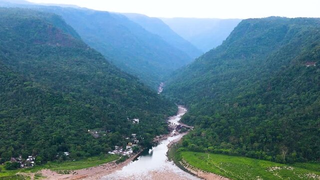 forested river through hill valley with small indigenous homes below. aerial