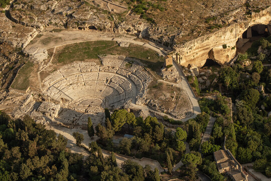 Aerial view of the ancient Greek Theatre of Syracuse nestled within the Neapolis Archaeological Park surrounded by lush trees and rocky terrain in Syracuse, Sicily, Italy.