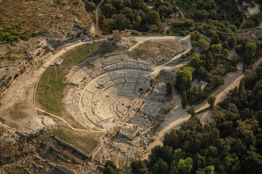 Aerial view of the ancient Greek Theatre of Syracuse, a historic stone amphitheater within the Neapolis Archaeological Park surrounded by trees and rocky terrain. Syracuse, Sicily, Italy.