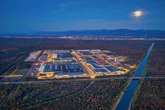 Aerial view of a large illuminated industrial complex surrounded by dense forest and a canal under a moonlit night sky in Mulhouse, Grand Est, France.