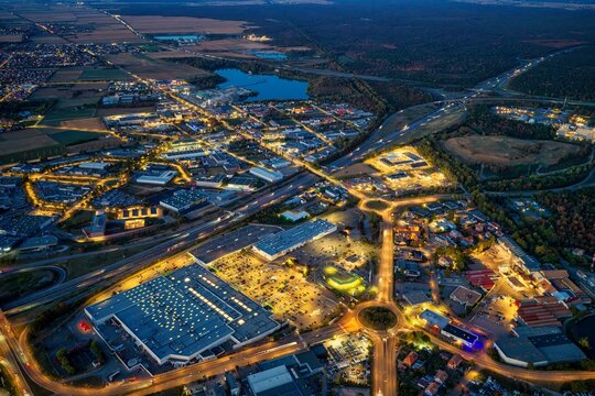 Aerial view of the illuminated industrial and commercial district of Mulhouse at night, featuring large warehouses, parking lots, road networks, and a nearby lake in Mulhouse, Grand Est, France.