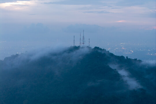 Aerial view of telecommunication towers atop a forested hill shrouded in mist during the late evening with distant city lights visible in Bukit Mertajam, Penang, Malaysia.