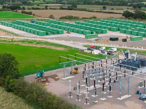 Aerial view of a large-scale battery energy storage system with rows of green container units, electrical substation infrastructure, and construction equipment in the United Kingdom.