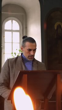 Man reading at church lectern with candle flames in foreground, standing beside arched window and religious icons while speaking or reciting inside quiet church interior, focused on text before him