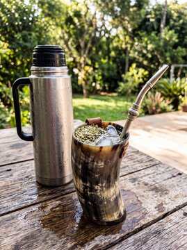 Traditional terere cold yerba mate drink in horn cup with metal straw and ice beside thermos on rustic wooden table outdoors