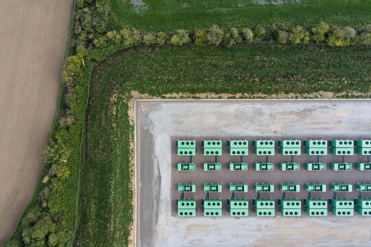 Aerial view of a large-scale battery energy storage system with rows of green battery units arranged on a gravel pad, surrounded by rural fields and hedgerows in the United Kingdom.