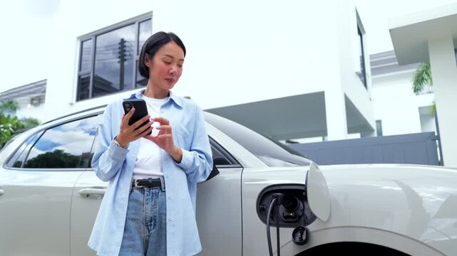Asian female using a smartphone app while charging her electric car at home. Smart EV home charging, green energy, and sustainable lifestyle.