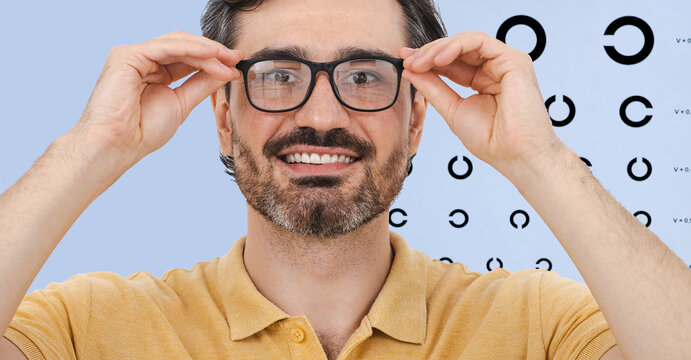 Adult man with beard smiling and adjusting his black glasses frame in front of a Landolt C eye test chart during a professional ophthalmology consultation.
