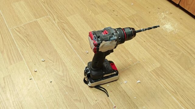 Cordless drill stands ready on wooden flooring for floor repair. Drill bit faces worn patch, and loose dust around boards suggests ongoing home fix work and careful drilling with battery powered tool.