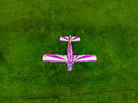 Aerial view of a small biplane with purple and white stripes parked on a vibrant green grass field in Latvia.