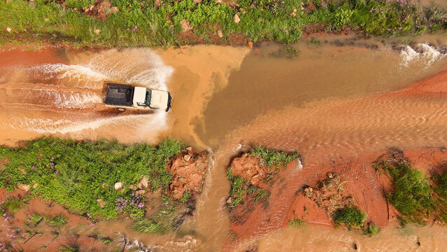 Aerial view of a 4x4 utility vehicle driving through a flooded dirt road, creating splashes in the muddy water, surrounded by green vegetation in Winterton, KwaZulu-Natal, South Africa.