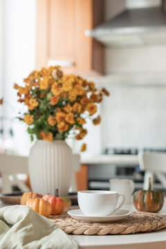 White tea cup with small pumpkins and orange flowers creates a relaxed autumn tea scene in a bright home kitchen. Vertical format.