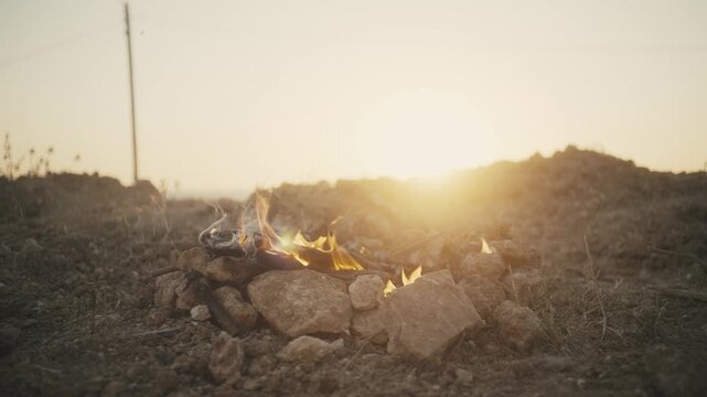 Campfire burning on rocks outdoors in nature during sunset with a person throwing wood