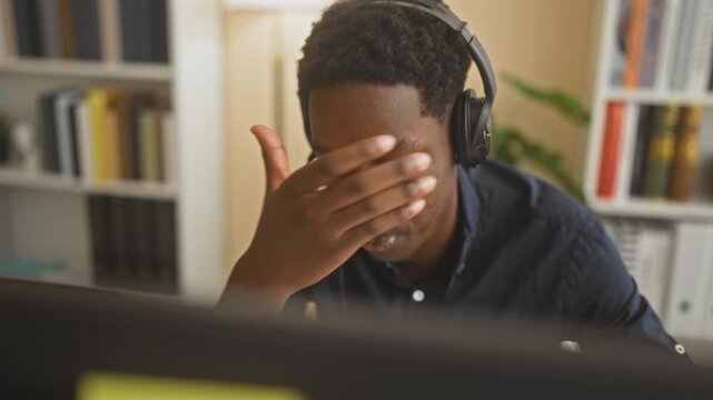 Young man wearing a headset covers his face with one hand and rests chin on fist during a call at office building; stress concentration.