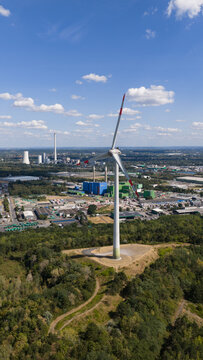 Aerial view of the wind turbine at Halde Hoppenbruch surrounded by lush forest with an industrial cityscape in the background in Herten, North Rhine-Westphalia, Germany.