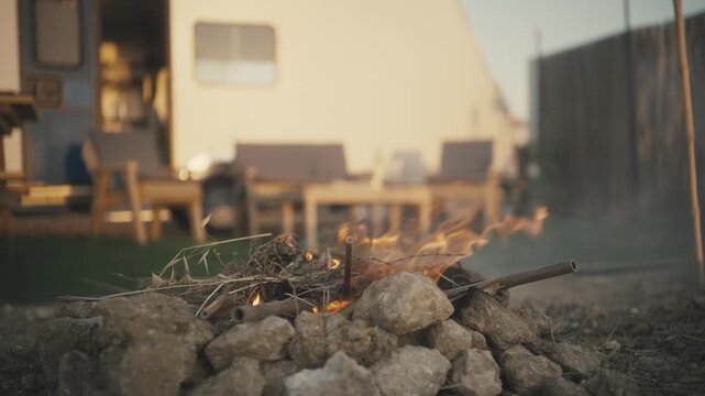 Small campfire burning on rocks outdoors with a camper van and outdoor furniture in the background
