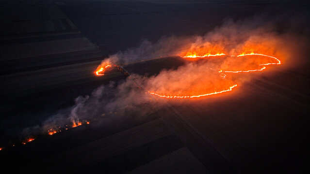 Aerial view of a wildfire spreading across agricultural fields with bright orange flames and thick smoke illuminating the dark landscape in Crepaja, Vojvodina, Serbia.