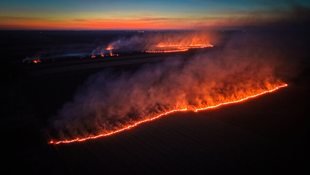 Aerial view of a wildfire spreading across agricultural fields at dusk, with bright orange flames and thick smoke plumes rising into the dark sky, Crepaja, Vojvodina, Serbia.