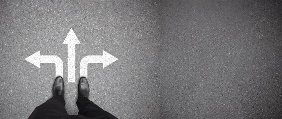 Fototapeta premium Top view of businessman standing on asphalt road with white painted direction arrows pointing left straight and right, conceptual image for business decision making and career path planning with