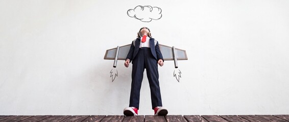 Fototapeta premium Ambitious young child in a formal business suit wearing aviator goggles and cardboard jetpack wings, looking up at hand-drawn clouds from a low angle perspective, symbolizing future career goals.