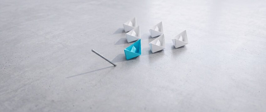 Blue paper boat leading a group of white origami boats on a grey stone surface, representing leadership, vision, and a shift in perspective for business success and strategic innovation.