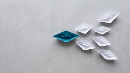 Fototapeta premium Top view of a unique blue paper boat leading a fleet of white boats on a grey textured background, conceptualizing leadership, business direction, teamwork, and standing out from the crowd.