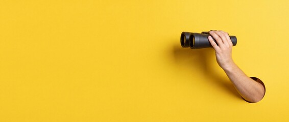 Fototapeta premium Hand holding black binoculars appearing through a round hole in a vibrant yellow background with wide copy space, representing discovery, business vision, and looking for new opportunities.