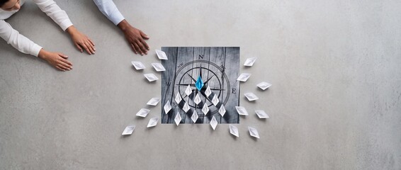 Fototapeta premium Business Leadership and Strategic Direction Concept Featuring a Blue Paper Boat Leading a Fleet of White Boats Over a Compass Graphic with Diverse People's Hands in a Wide Top View