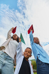 Naklejka premium Sisters raising paper airplanes with younger brother in park with excited smiles. Creative learning, craft supplies, afterschool programs concept.